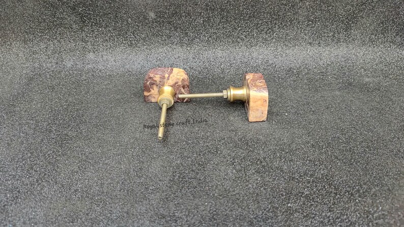 May include: Two decorative cabinet knobs. Each knob features a rectangular, brown and tan stone face with a brass stem and hardware. The knobs are designed for furniture and home decor.