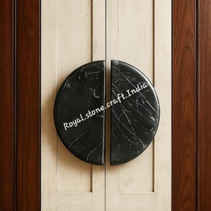 Op de afbeelding: Een paar zwarte marmeren kastknoppen, elk een halve cirkel, gemonteerd op een crèmekleurige kastdeur. De knoppen hebben witte adering en de tekst "Royal.stone.craft.India". De kast wordt geflankeerd door donkere houten panelen.