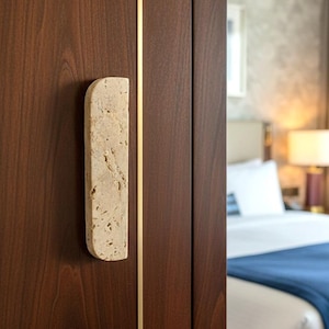May include: Close-up of a dark brown wooden cabinet door with a rectangular, light-colored stone handle. The door has gold trim. A bed with white linens and a blue blanket is visible in the background.