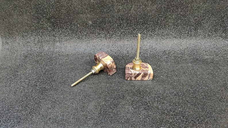 May include: Two decorative cabinet knobs with a brown and beige marbled stone design and brass hardware. One knob is lying on its side, while the other is upright. The text "i.India" is visible on the base of the upright knob.