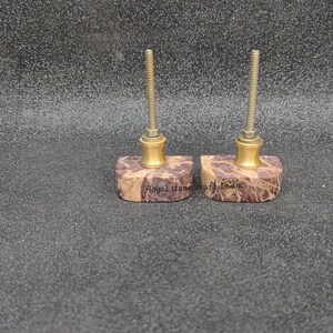 May include: Two decorative cabinet knobs. Each knob features a rectangular, patterned stone base with a brass-colored metal accent and a threaded rod. The stone base has a mix of brown, beige, and purple hues. The text "Royal Stone of India" is visible on the base.