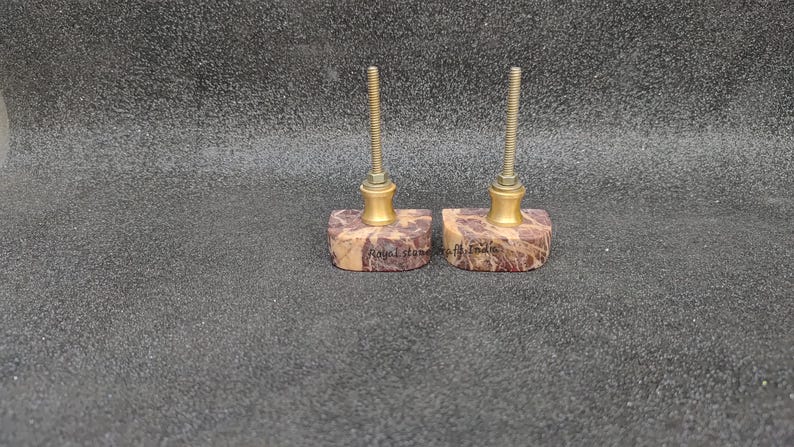 May include: Two decorative cabinet knobs. Each knob features a rectangular, patterned stone base with a brass-colored metal accent and a threaded rod. The stone base has a mix of brown, beige, and purple hues. The text "Royal Stone of India" is visible on the base.