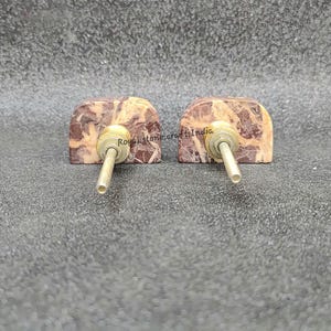 May include: Two decorative cabinet knobs. Each knob features a rectangular, arch-shaped stone with a mix of brown and beige tones. A brass-colored metal rod extends from the center of each stone, designed for easy installation. These knobs are ideal for adding a touch of natural elegance to furniture.