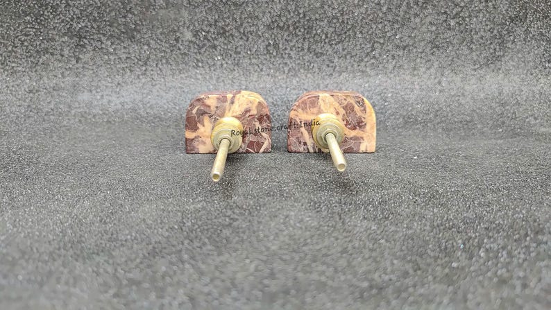 May include: Two decorative cabinet knobs. Each knob features a rectangular, arch-shaped stone with a mix of brown and beige tones. A brass-colored metal rod extends from the center of each stone, designed for easy installation. These knobs are ideal for adding a touch of natural elegance to furniture.