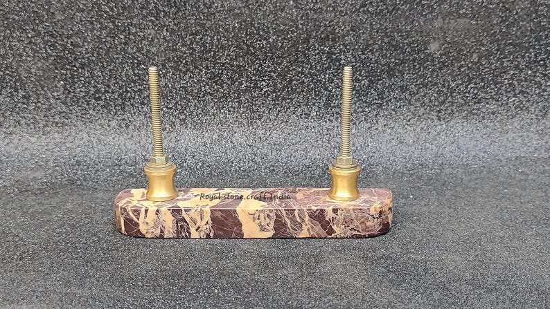 May include: A marble and brass pen holder with two threaded posts. The rectangular base features brown and tan veining. The text "Royal stone craft India" is visible on the base.