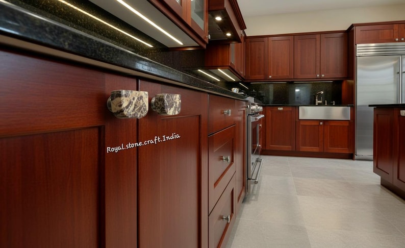 May include: A kitchen interior with dark wood cabinetry and a black countertop. Two decorative marble cabinet pulls are visible. The text "Royal.stone.craft.India" is printed on the cabinet. Stainless steel appliances and a light-colored floor complete the scene.