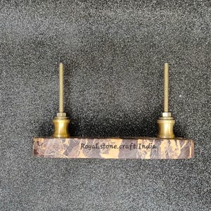 May include: A rectangular cabinet pull with two brass-colored posts and a marble-patterned base. The text "Royal stone.craft.India" is printed on the base. The hardware is set against a textured gray background.