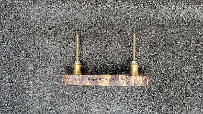 May include: A rectangular cabinet pull with two brass-colored posts and a marble-patterned base. The text "Royal stone.craft.India" is printed on the base. The hardware is set against a textured gray background.