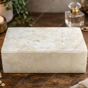 May include: A rectangular, off-white marble box with a lid. The box is on a wooden surface, with a lit candle, jewelry, and a perfume bottle in the background. The box is approximately 8 inches long.