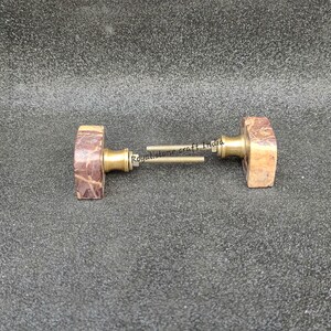 May include: Two rectangular door knobs with a brown and tan marbled appearance. The knobs have a brass-colored base and are connected by a metal rod. The knobs are likely for a cabinet or door.