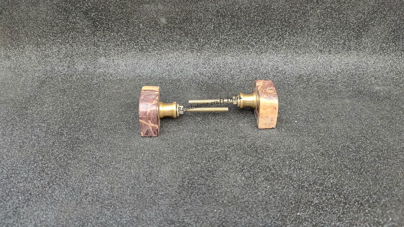 May include: Two rectangular door knobs with a brown and tan marbled appearance. The knobs have a brass-colored base and are connected by a metal rod. The knobs are likely for a cabinet or door.