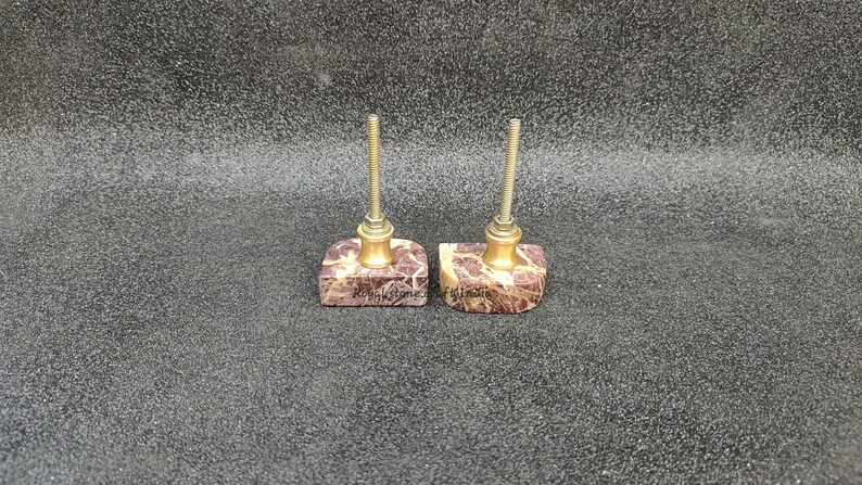 May include: Two decorative cabinet knobs. Each knob features a square, patterned stone base with a brass-colored metal accent and a threaded post. The stone base has a brown and white pattern. The knobs are for furniture.