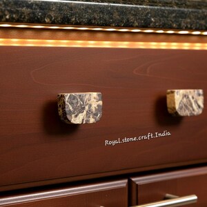 May include: Two square cabinet knobs made of stone with a marbled pattern of gray, brown, and beige. The knobs are mounted on a dark brown cabinet door. The text "Royal.stone.craft.India" is visible.