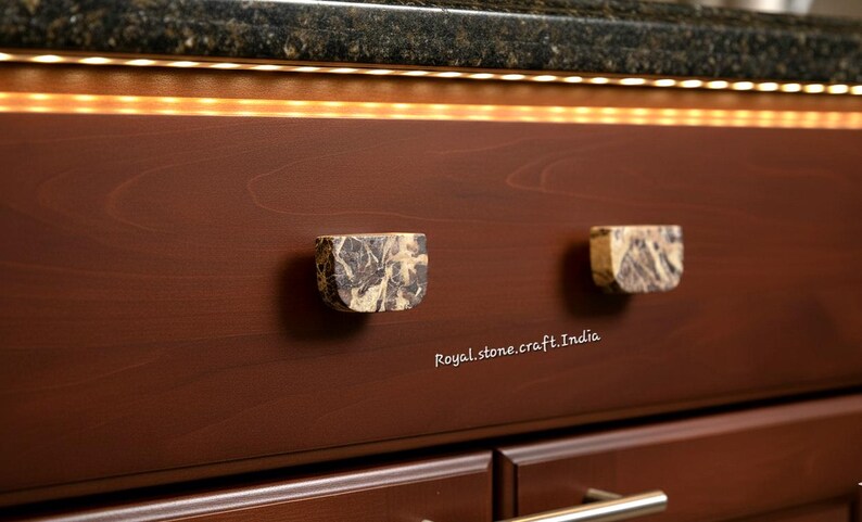 May include: Two square cabinet knobs made of stone with a marbled pattern of gray, brown, and beige. The knobs are mounted on a dark brown cabinet door. The text "Royal.stone.craft.India" is visible.