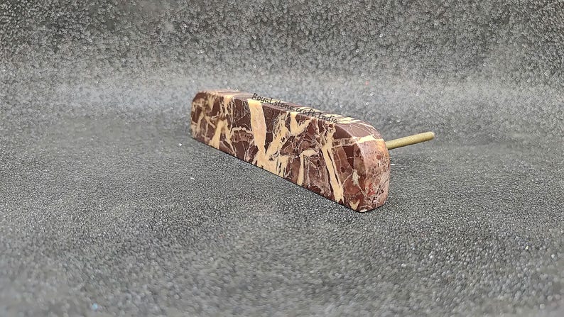 May include: A rectangular cabinet pull with a marble-like pattern in shades of brown and cream. The pull has a brass-colored stem extending from one end. The text "Royal Stone Craft India" is visible on the pull.
