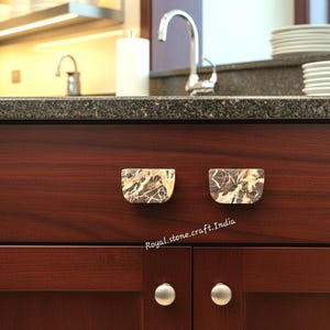Könnte beinhalten: Nahaufnahme eines dunklen Holzschranks mit zwei marmorähnlichen Schubladengriffen. Die Griffe sind hellbeige mit dunkelbraunen Maserungen. Die Schranktüren haben silberne Knöpfe. Der Hintergrund zeigt eine Küche mit Spüle und Arbeitsplatte.