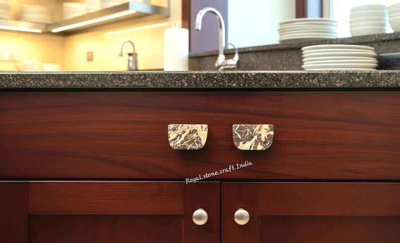 May include: Close-up of a dark wood cabinet with two marble-like drawer pulls. The pulls are a light beige color with dark brown veining. The cabinet doors have silver knobs. The background shows a kitchen with a sink and countertop.