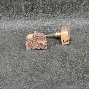May include: Two decorative cabinet knobs with a natural stone appearance. The knobs have a square shape with a reddish-brown and beige marbled pattern. The hardware is brass-colored. The text "Royal stone craft India" is visible on the knobs.