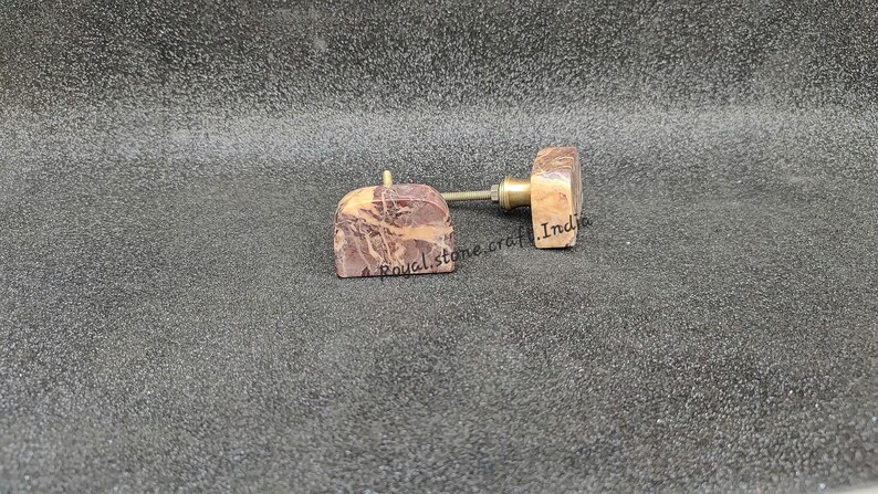May include: Two decorative cabinet knobs with a natural stone appearance. The knobs have a square shape with a reddish-brown and beige marbled pattern. The hardware is brass-colored. The text "Royal stone craft India" is visible on the knobs.