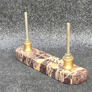 May include: A marble pen holder with two brass posts. The rectangular base features brown and tan veining. The posts are threaded and have a decorative brass base. The text "Royal Stone Crafts India" is visible on the base.
