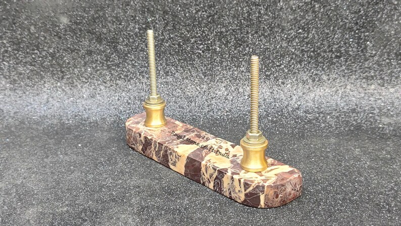 May include: A marble pen holder with two brass posts. The rectangular base features brown and tan veining. The posts are threaded and have a decorative brass base. The text "Royal Stone Crafts India" is visible on the base.