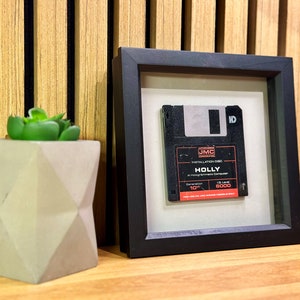 May include: A black framed picture containing a vintage floppy disk. The floppy disk is labelled "HOLLY" and "JMC Installation Disc".
