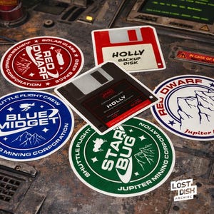 May include: A selection of circular and square stickers with a science fiction theme. The stickers display text such as "Red Dwarf", "Blue Midget", and "Star Bug". One sticker is a floppy disk labelled "Holly Backup Disk".