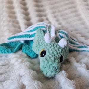 May include: A handmade crochet dragon in shades of teal and white. The dragon has large, black eyes with gold accents and is lying on a soft, white, textured surface. The wings are teal with white stripes.