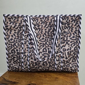 以下が含まれることがあります： 茶色と黒のヒョウ柄の長方形トートバッグ。バッグには白と黒のストライプのハンドルとトリムが付いています。バッグは木製の表面に置かれています。