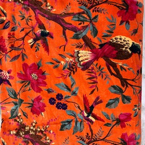 以下が含まれることがあります： 鮮やかなオレンジ色の生地に、花と鳥の模様が描かれています。赤い花、青緑色の葉、茶色の枝に、飛んでいる鳥がデザインされています。この生地は柔らかくビロードのような質感で、室内装飾や装飾プロジェクトに最適です。