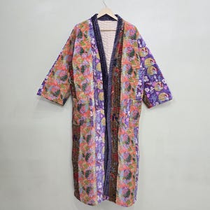 Könnte beinhalten: Langer, offen geschnittener Morgenmantel mit Patchwork-Design. Der Morgenmantel weist verschiedene Blumenmuster in Lila, Rosa, Orange und Grün auf. Der Morgenmantel hängt an einem Holzbügel vor einem schlichten Hintergrund.