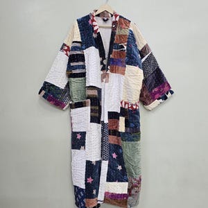 Op de afbeelding: Een lange patchwork badjas met een gewatteerd ontwerp. De badjas heeft een verscheidenheid aan stoffen in tinten blauw, wit, roze en groen. De badjas hangt aan een houten hanger tegen een neutrale achtergrond.