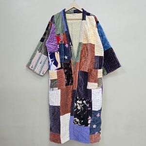Könnte beinhalten: Ein Kimono-ähnlicher Patchwork-Mantel in verschiedenen Farben, darunter Lila, Blau, Braun und Grün. Der Mantel besteht aus verschiedenen zusammengenähten Stoffquadraten. Er hängt an einem Holzbügel vor einem neutralen Hintergrund.