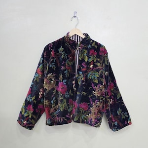 Peut inclure: Veste en velours bleu marine foncé avec un motif floral vibrant dans les tons de rose, vert et violet. La veste présente un col mandarin et une doublure rayée. Elle est suspendue à un cintre en bois.