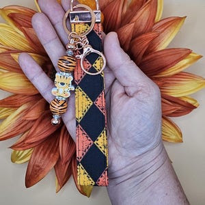 May include: A black and orange checkered fabric keychain with a gold metal ring and a tiger bead charm. The charm has a white face and orange stripes.