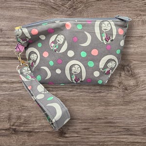 May include: A gray fabric zippered pouch with a black and white print of a cartoon character. The pouch has a gray fabric strap with a black and white print of a crescent moon.