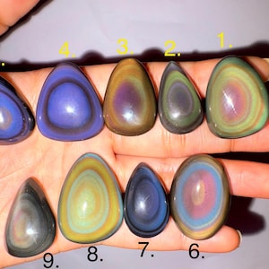 May include: Nine polished, oval-shaped obsidian stones in various colors, including blue, green, yellow, and purple. Each stone has a unique pattern of concentric circles, creating a striking visual effect. The stones are displayed on a hand.