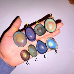 May include: A collection of eight polished rainbow obsidian stones in various shapes, including oval, teardrop, and round. The stones display iridescent colors like blue, green, yellow, and purple, with concentric patterns. The stones are held in a hand.