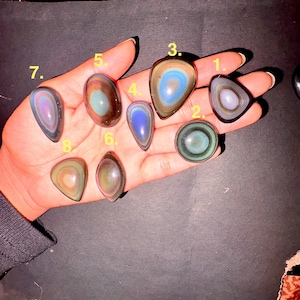 May include: A hand displays eight polished obsidian stones in various shapes. The stones feature iridescent bands of color, including blues, greens, purples, and browns. The stones are oval, teardrop, and triangular shapes.
