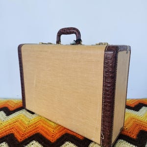 May include: A vintage tan suitcase with brown leather trim and handle. The suitcase features brass-coloured hardware and a textured canvas exterior. The corners and handle are a darker brown, contrasting the tan. The rectangular case appears to be in good condition.