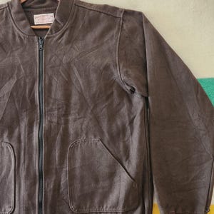 Peut inclure: Veste de bombardier marron avec une fermeture &eacute;clair int&eacute;grale, deux poches avant et des poignets c&ocirc;tel&eacute;s. La veste est suspendue &agrave; un cintre en bois. Le tissu semble &ecirc;tre un mat&eacute;riau doux et textur&eacute;.