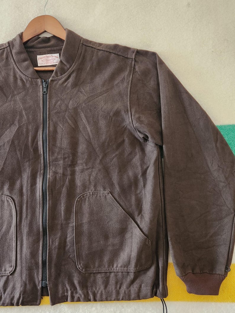 Peut inclure: Veste de bombardier marron avec une fermeture &eacute;clair int&eacute;grale, deux poches avant et des poignets c&ocirc;tel&eacute;s. La veste est suspendue &agrave; un cintre en bois. Le tissu semble &ecirc;tre un mat&eacute;riau doux et textur&eacute;.