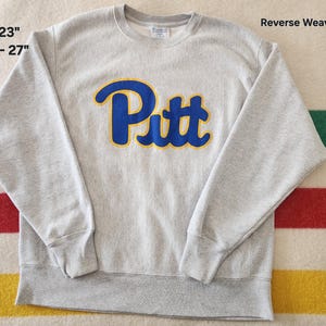 May include: A light gray crewneck sweatshirt with the word "Pitt" in blue letters outlined in yellow. The sweatshirt is 23 inches across the chest and 27 inches long. The text "Reverse Weave" is visible.