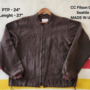 Peut inclure: Veste de bombardier marron avec fermeture &eacute;clair compl&egrave;te, deux poches avant et poignets c&ocirc;tel&eacute;s. La veste porte l'inscription "CC Filson Co. Seattle MADE IN USA". La veste mesure 61 cm de PTP et 69 cm de longueur.