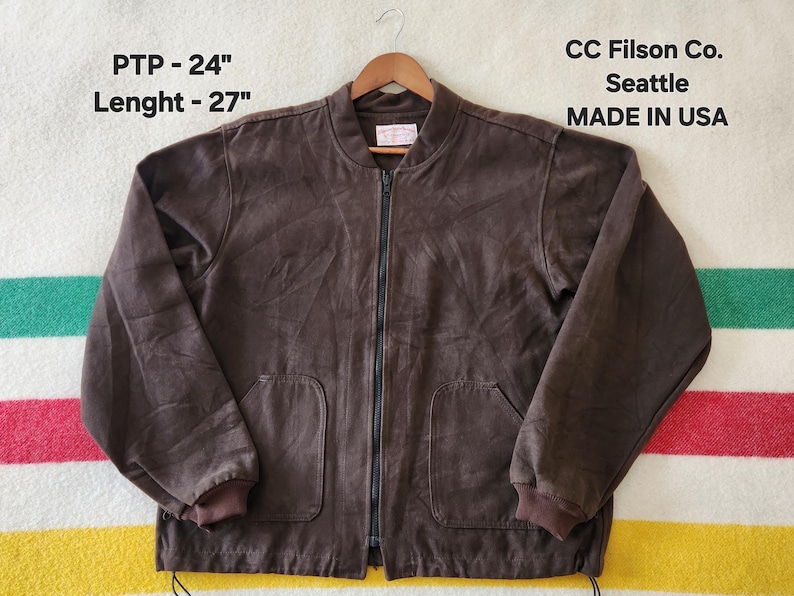 Peut inclure: Veste de bombardier marron avec fermeture &eacute;clair compl&egrave;te, deux poches avant et poignets c&ocirc;tel&eacute;s. La veste porte l'inscription "CC Filson Co. Seattle MADE IN USA". La veste mesure 61 cm de PTP et 69 cm de longueur.
