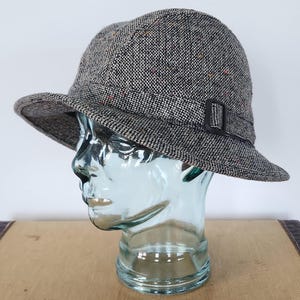 Peut inclure: Chapeau cloche en tweed gris avec un petit bord et une boucle noire. Le chapeau présente des mouchetures de couleur et est présenté sur une tête en verre. Style classique.