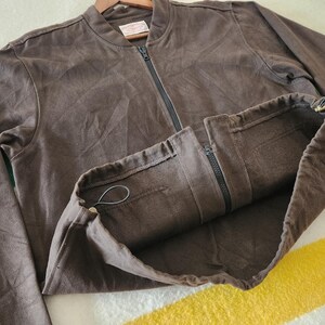 Peut inclure: Veste de bombardier marron avec une fermeture &eacute;clair sur toute la longueur et un ourlet &agrave; cordon de serrage. La veste est faite d'une mati&egrave;re douce et a une coupe d&eacute;contract&eacute;e. La veste est pr&eacute;sent&eacute;e sur un cintre en bois.