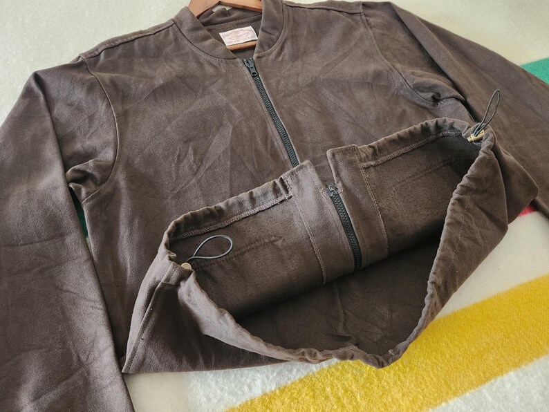 Peut inclure: Veste de bombardier marron avec une fermeture &eacute;clair sur toute la longueur et un ourlet &agrave; cordon de serrage. La veste est faite d'une mati&egrave;re douce et a une coupe d&eacute;contract&eacute;e. La veste est pr&eacute;sent&eacute;e sur un cintre en bois.