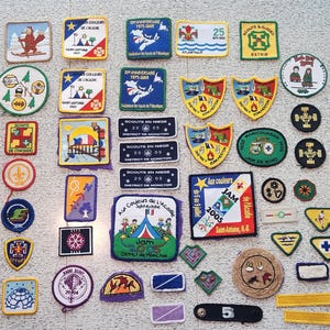 May include: A collection of 45 vintage scout patches in various shapes and colors. The patches feature text, illustrations, and logos, including the text "LOT OF 45", "JAM 2005", and "30 ANNIVERSAIRE 1975-2005".