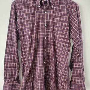 Könnte beinhalten: Ein langärmeliges Button-Down-Hemd mit einem Karomuster in Burgunderrot, Marineblau und Creme. Das Hemd hängt an einem Holzbügel. Es hat einen Button-Down-Kragen und weiße Knöpfe.